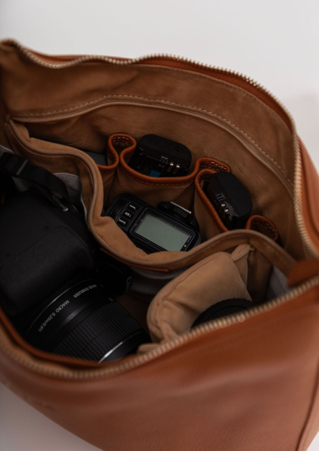 A tan leather camera bag shown open, filled with camera gear including a DSLR camera, lens, and small accessories, on a plain light background.