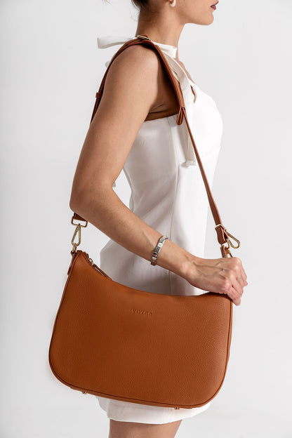 A woman in a white sleeveless dress carries a tan leather shoulder bag with a curved bottom and gold-tone hardware against a light gray studio background.