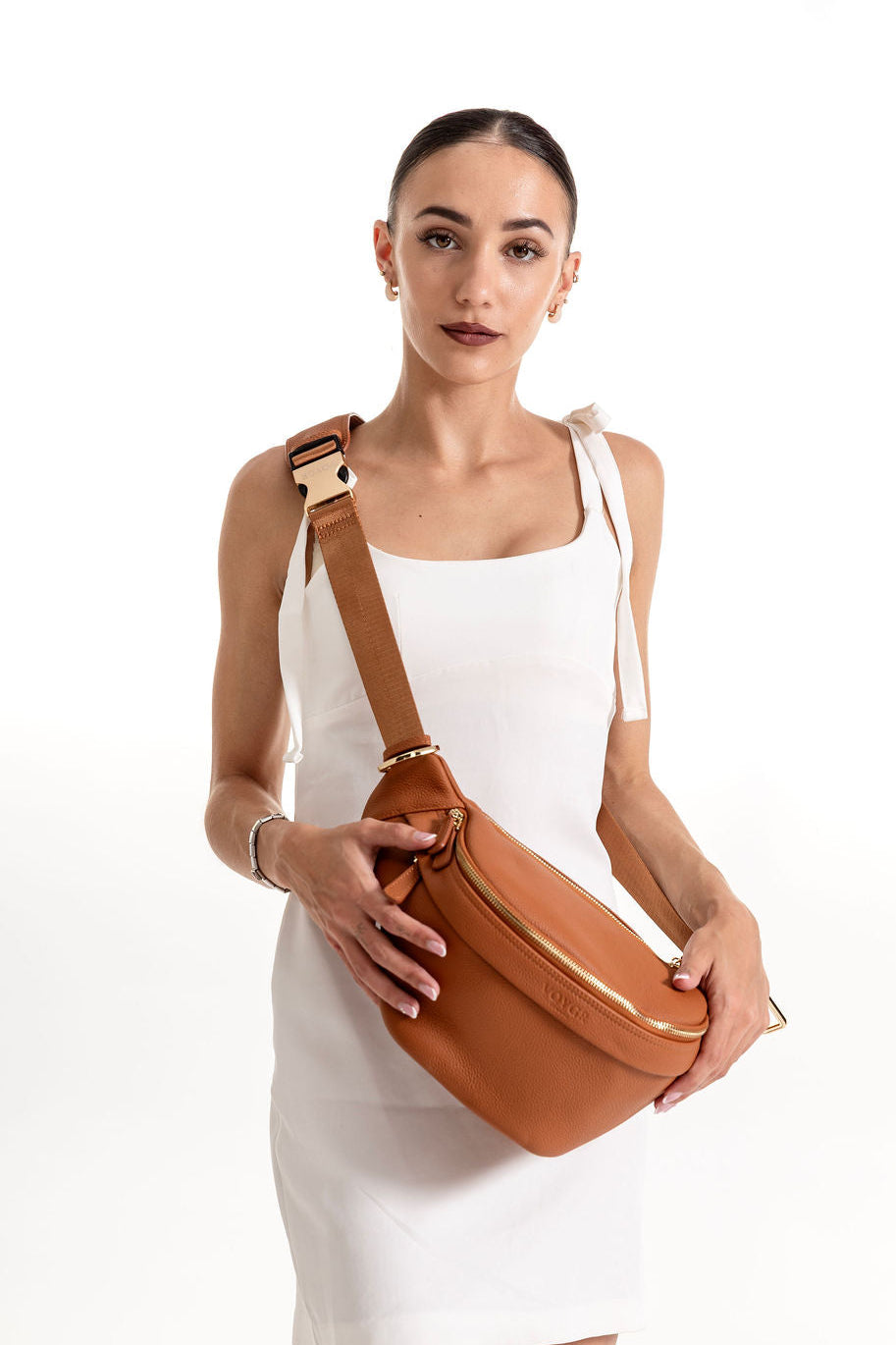 A woman wearing a white sleeveless dress models a tan leather crossbody bag with a gold-tone buckle and zipper, set against a plain white studio background.