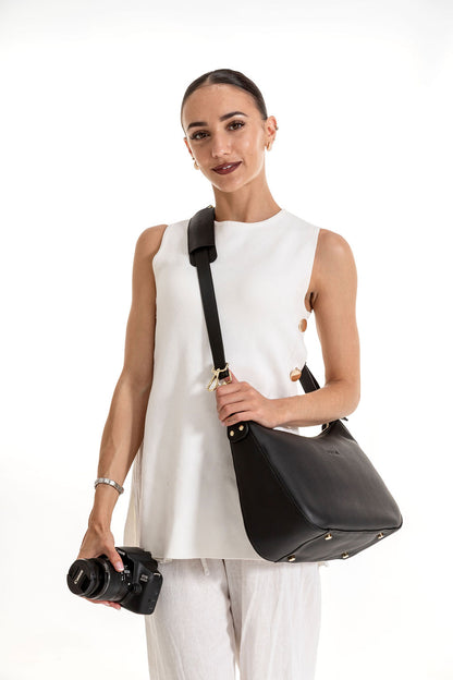 A woman wearing a white sleeveless top and white pants carries a black leather shoulder bag with gold-tone hardware. She is holding a black camera against a clean white studio background.