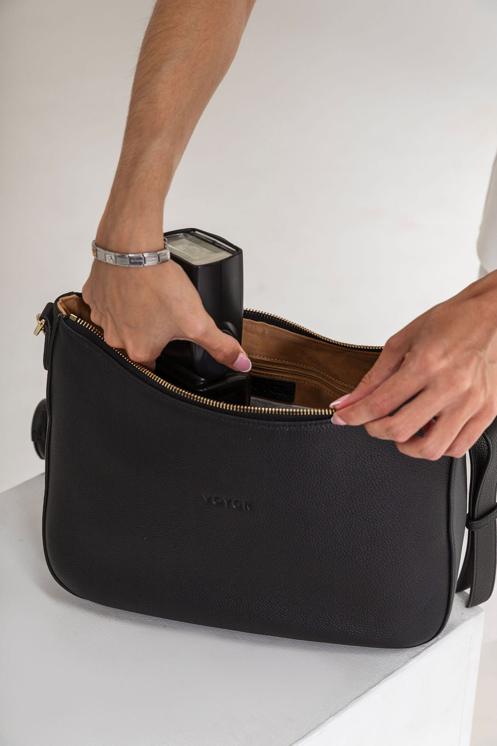A black leather shoulder bag with gold zipper details, shown with a person's hands and arms inserting a black camera into its interior. The brand name 'VOGUE' is debossed on the front.