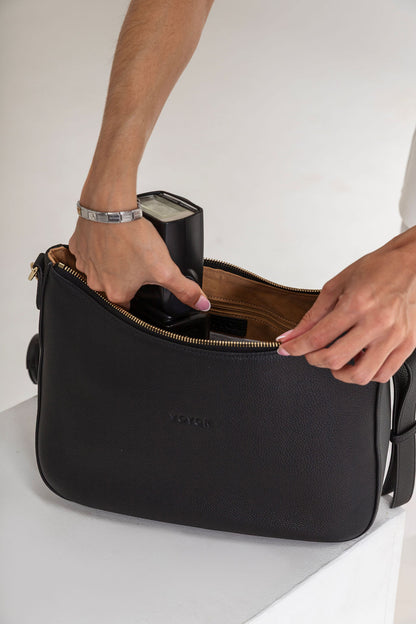 A black leather shoulder bag with gold zipper details, shown with a person's hands and arms inserting a black camera into its interior. The brand name 'VOGUE' is debossed on the front.