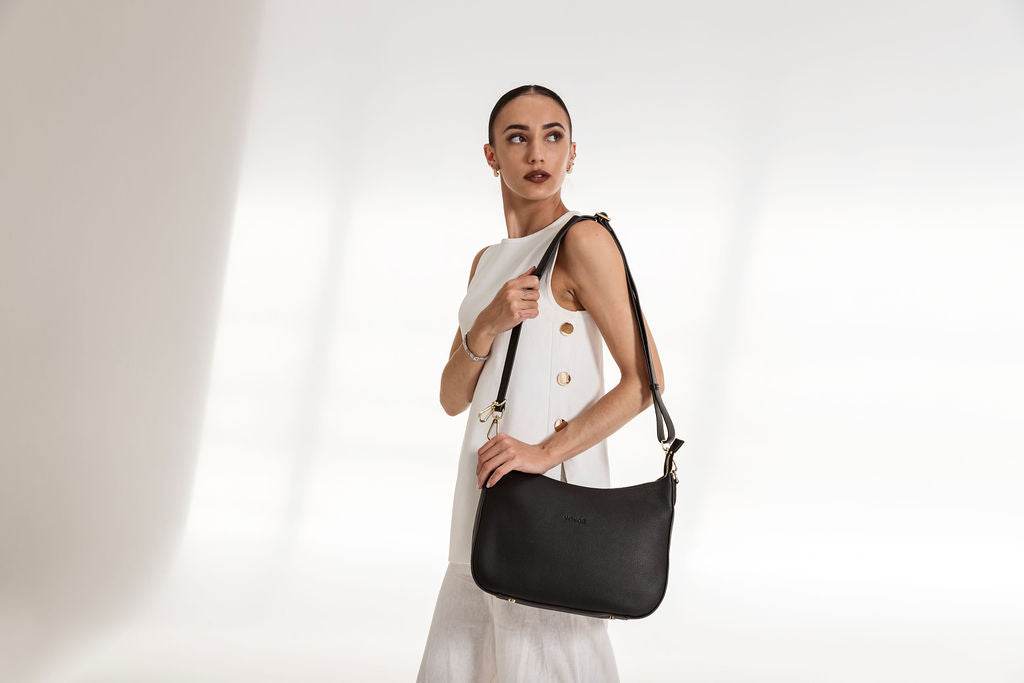 A woman wearing a white sleeveless dress with gold buttons carries a black leather shoulder bag with a gold-tone logo plate, set against a bright white studio background with soft diagonal shadows.