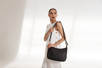 A woman wearing a white sleeveless dress with gold buttons carries a black leather shoulder bag with a gold-tone logo plate, set against a bright white studio background with soft diagonal shadows.