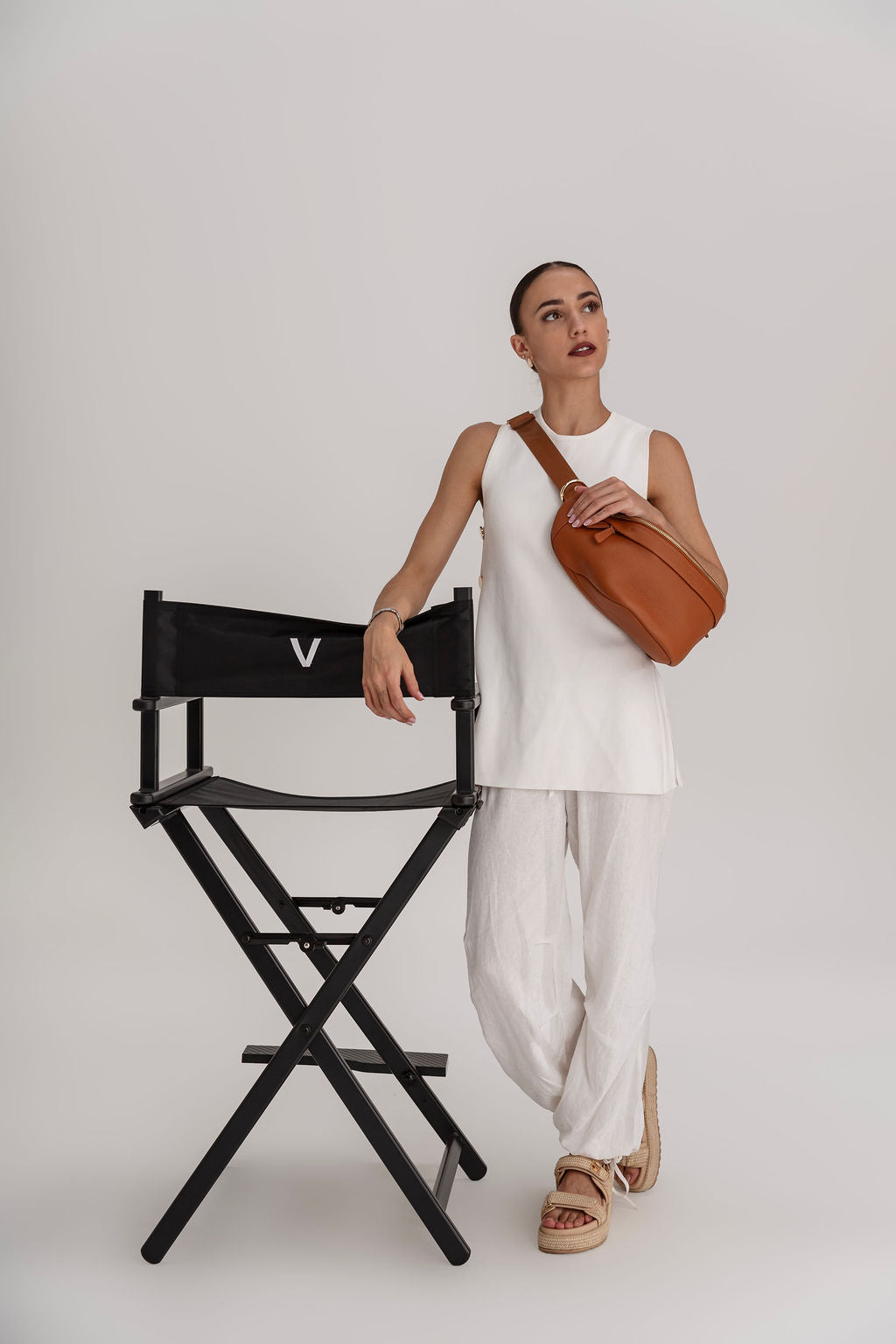 A woman models a brown leather crossbody bag with a wide strap, styled with a white sleeveless top and white pants, standing next to a black director's chair with a white 'V' logo.