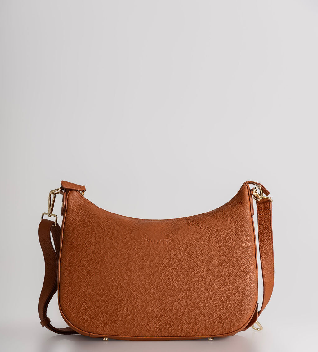A brown pebbled leather crescent-shaped shoulder bag with a matching strap and gold-tone hardware, photographed against a light gray studio background.