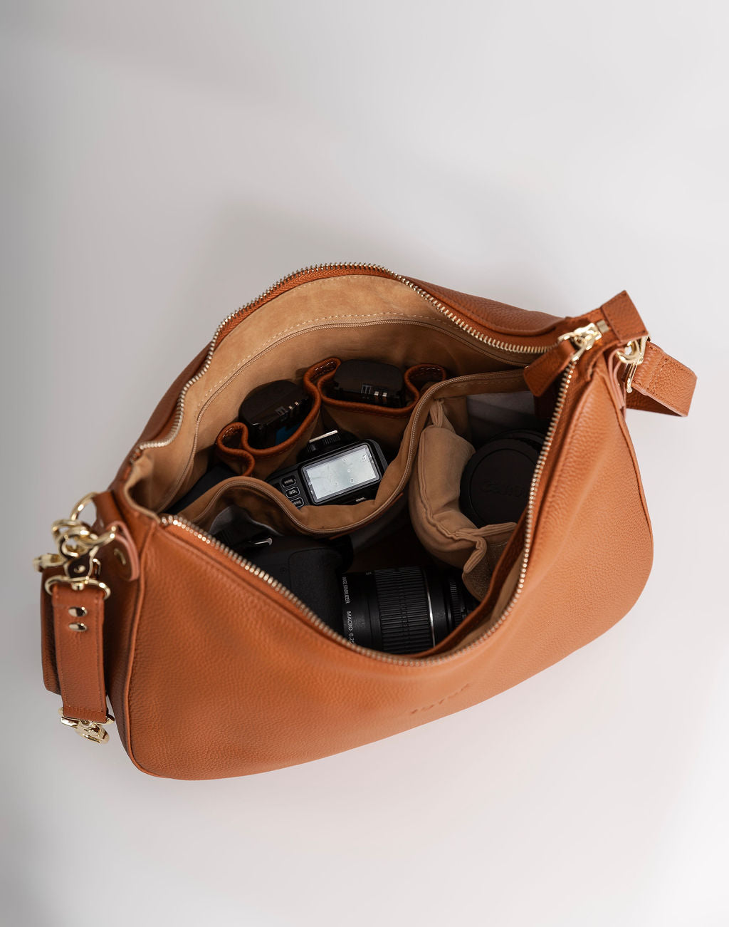 An open tan leather shoulder bag with gold-tone hardware, shown from above on a light gray background. The bag is filled with camera gear, including a camera body, lenses, and a strap.