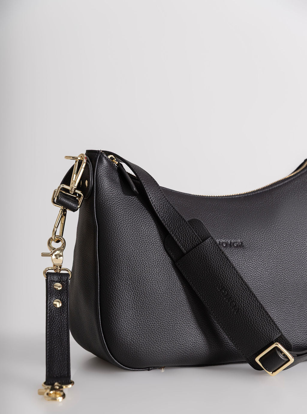 A black pebbled leather shoulder bag with a wide adjustable strap and gold-tone hardware, shown on a light gray studio background.