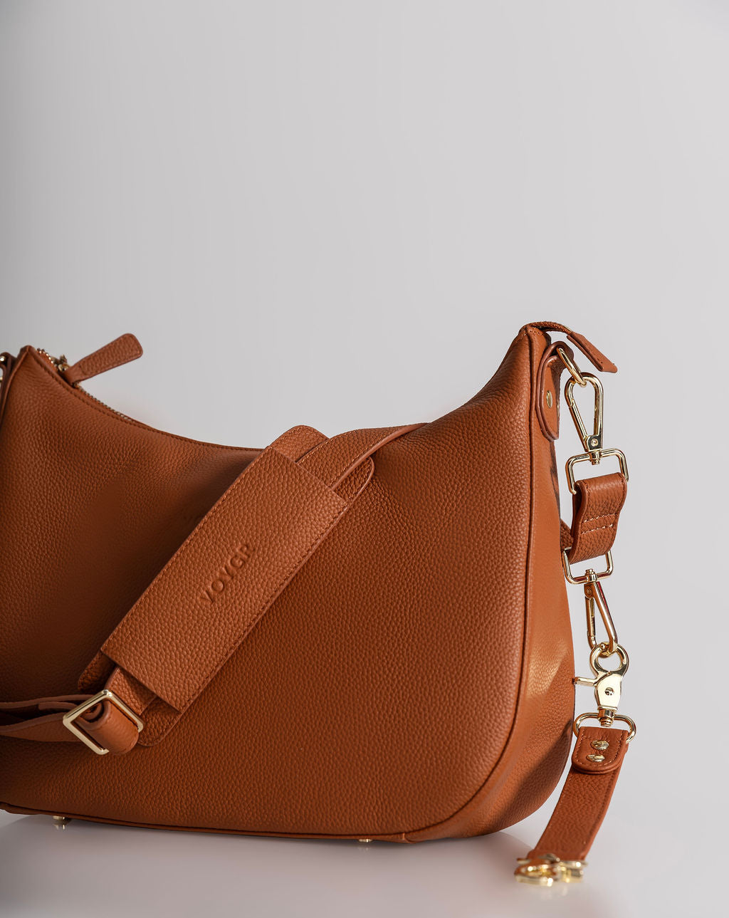 A brown pebbled leather shoulder bag with a wide adjustable strap and gold-tone hardware, photographed on a light gray studio background.