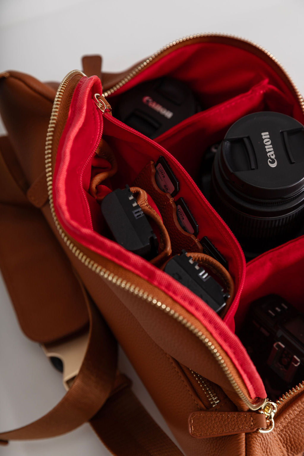 A close-up of a brown leather camera bag with a bright red interior organizer, shown open with a Canon lens and other camera accessories inside.