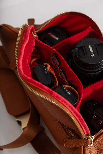 A close-up of a brown leather camera bag with a bright red interior organizer, shown open with a Canon lens and other camera accessories inside.